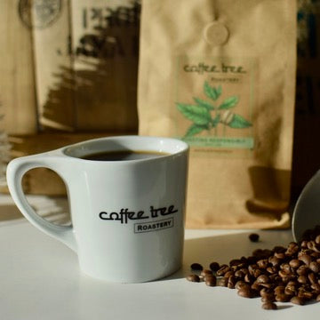 Coffee Tree Roastery mug filled with coffee beside roasted beans and coffee bag in warm natural light